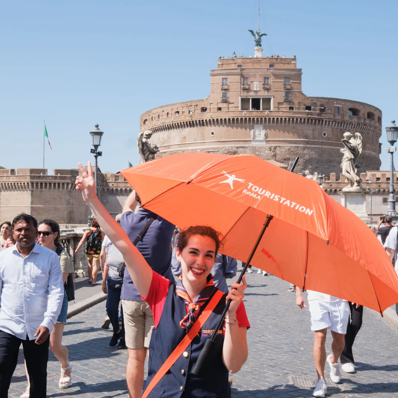 A cheerful woman holding an orange umbrella stands on a bridge with the Castel Sant'Angelo and tourists in the background.