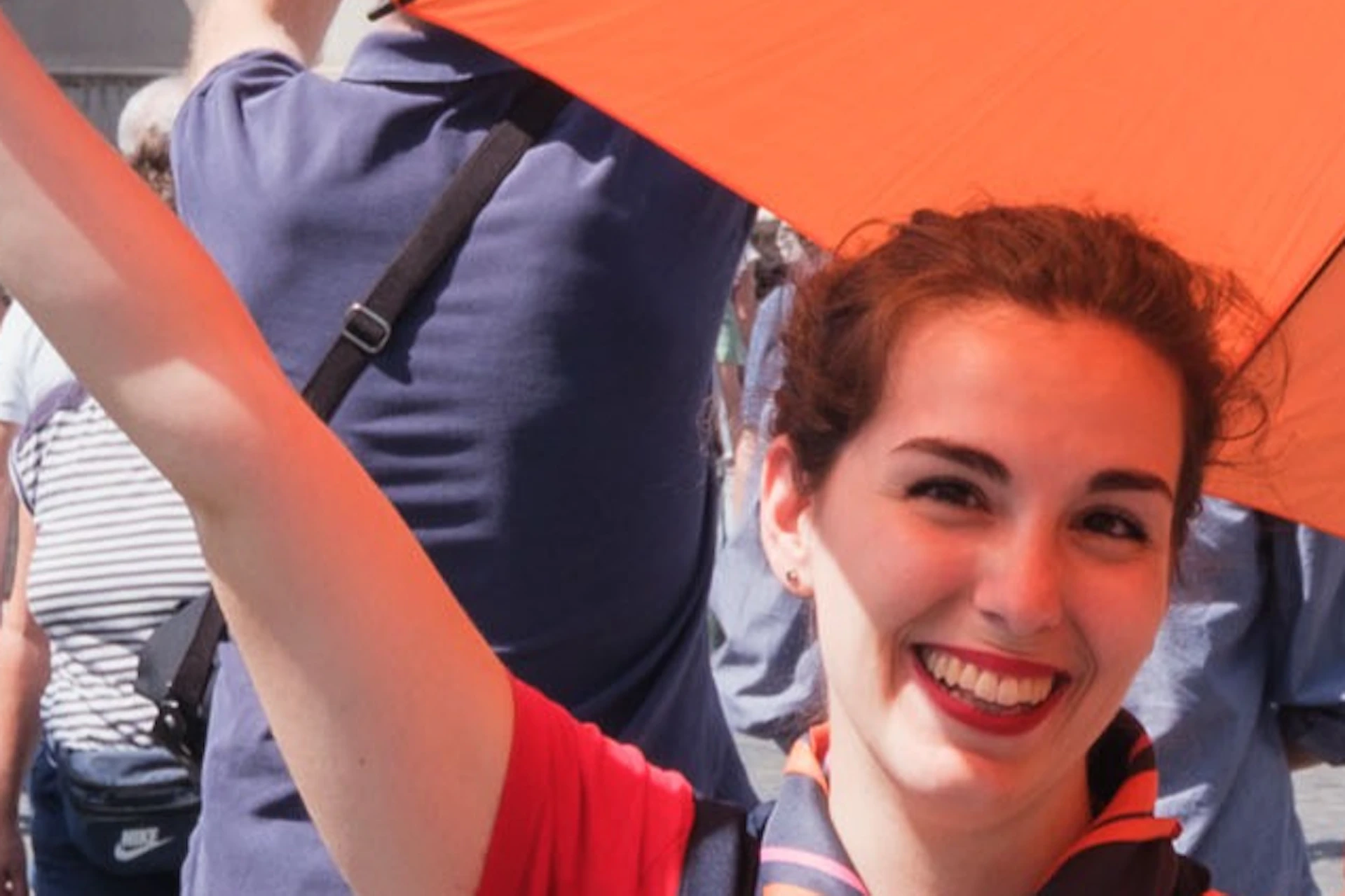 A cheerful woman holding an orange umbrella stands on a bridge with the Castel Sant'Angelo and tourists in the background.