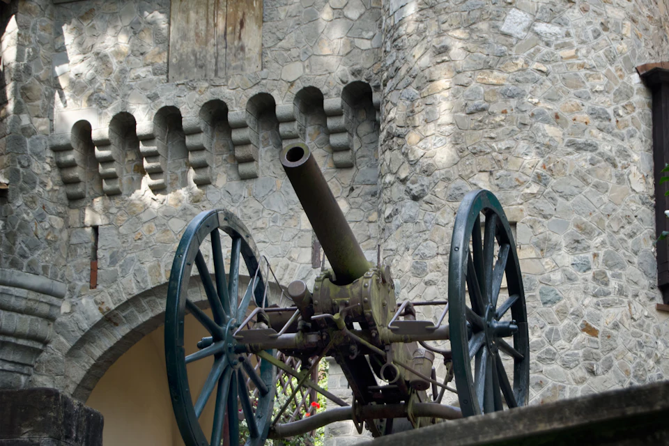 A vintage cannon with large wheels is positioned in front of a stone fortress wall with arched and rectangular openings.