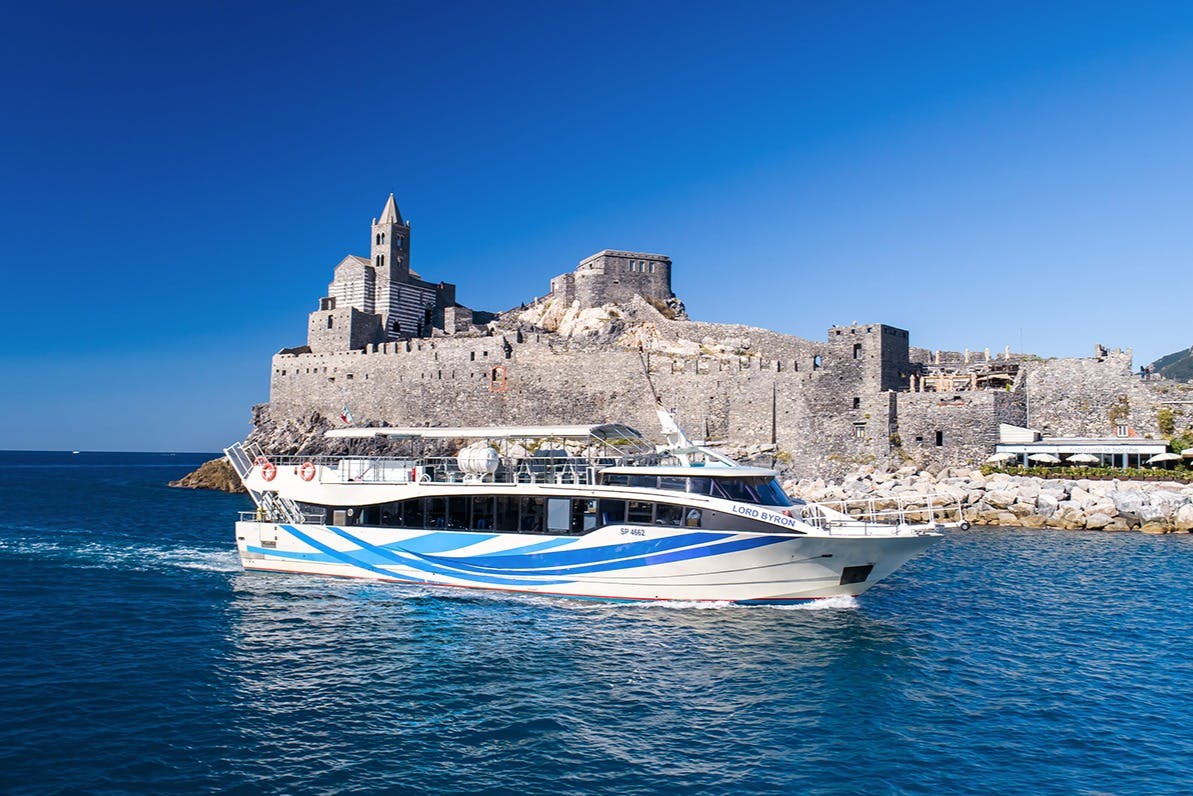 A white and blue tour boat on the water near a historic stone fortress under a clear blue sky.