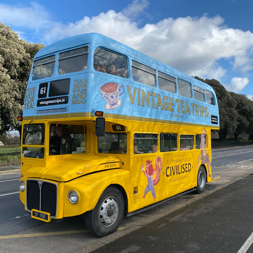 A bright yellow and blue double-decker bus, labeled "Vintage Tea Trips," is parked on a street near a park under a blue sky.