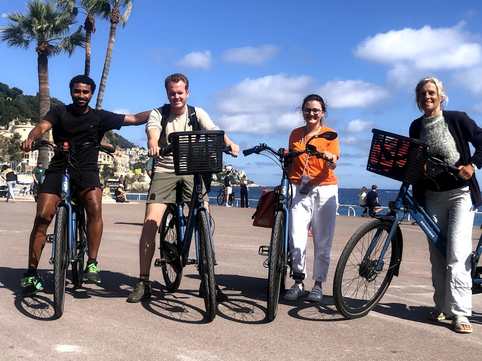 Three people stand with bicycles on a paved waterfront, with the ocean and partly cloudy sky in the background.