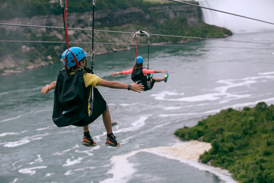 Two people wearing helmets and harnesses zip-lining over a river, with distant waterfalls and greenery in the background.