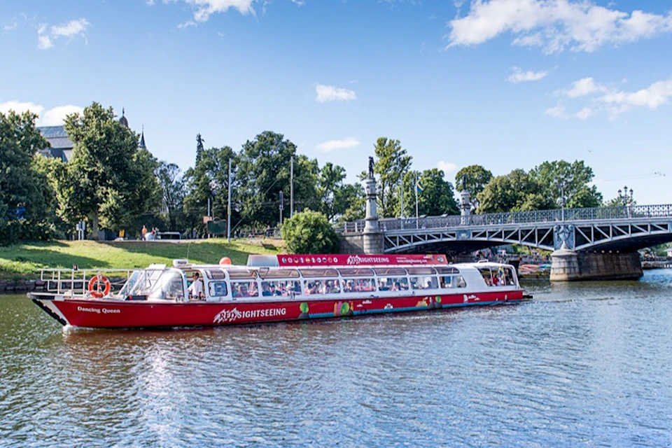 A red sightseeing boat cruises on a river with a lush green park and an ornate bridge in the background under a blue sky.