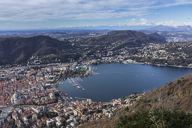 Вид с воздуха на город вокруг большого озера, окруженный холмами и горами, с лодками, пристыкованными к пристани, и зданиями по всей территории.