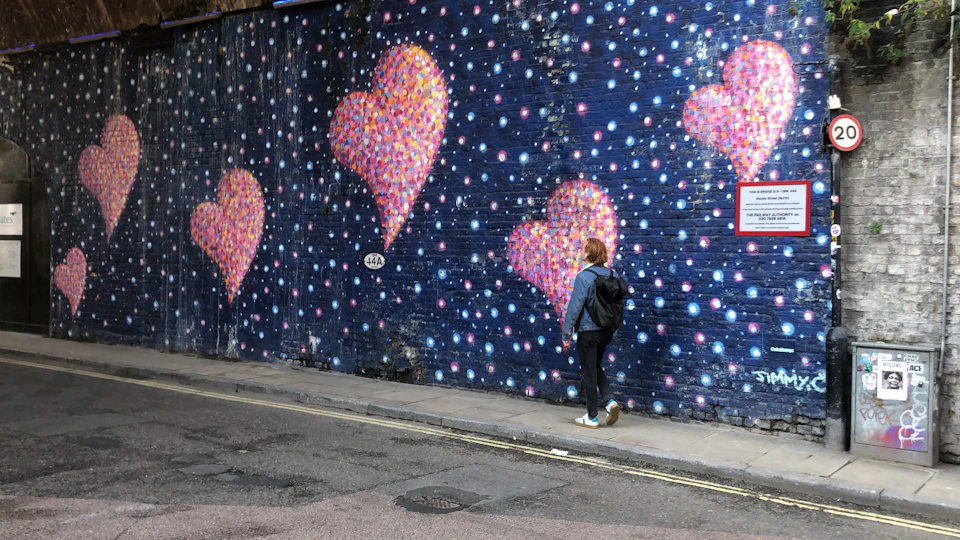 A person with a backpack walks past a mural of colorful, textured hearts and dots on a dark blue wall.