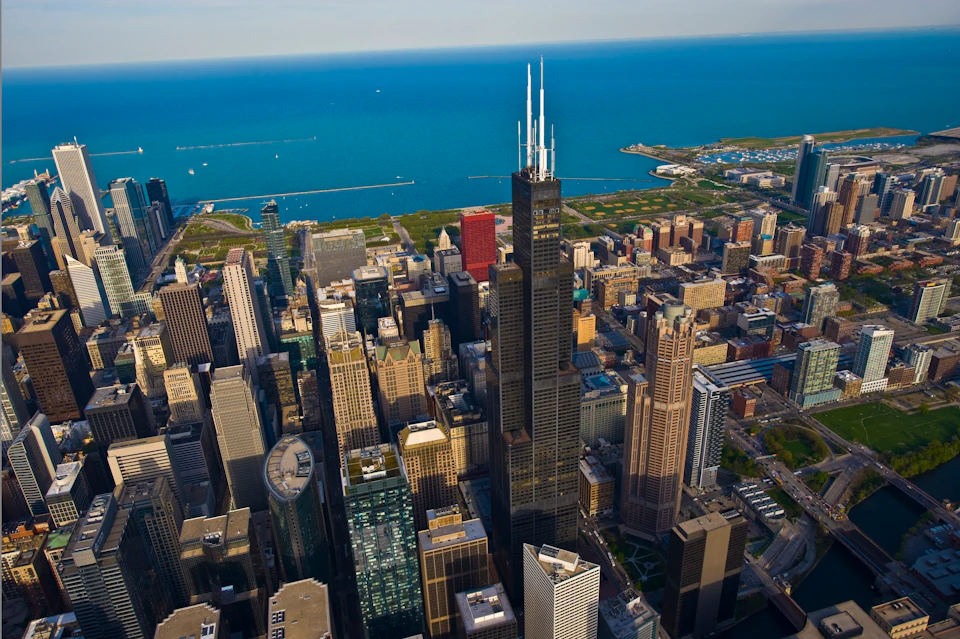 Aerial view of a cityscape with modern skyscrapers, green spaces, and a large body of water in the background.