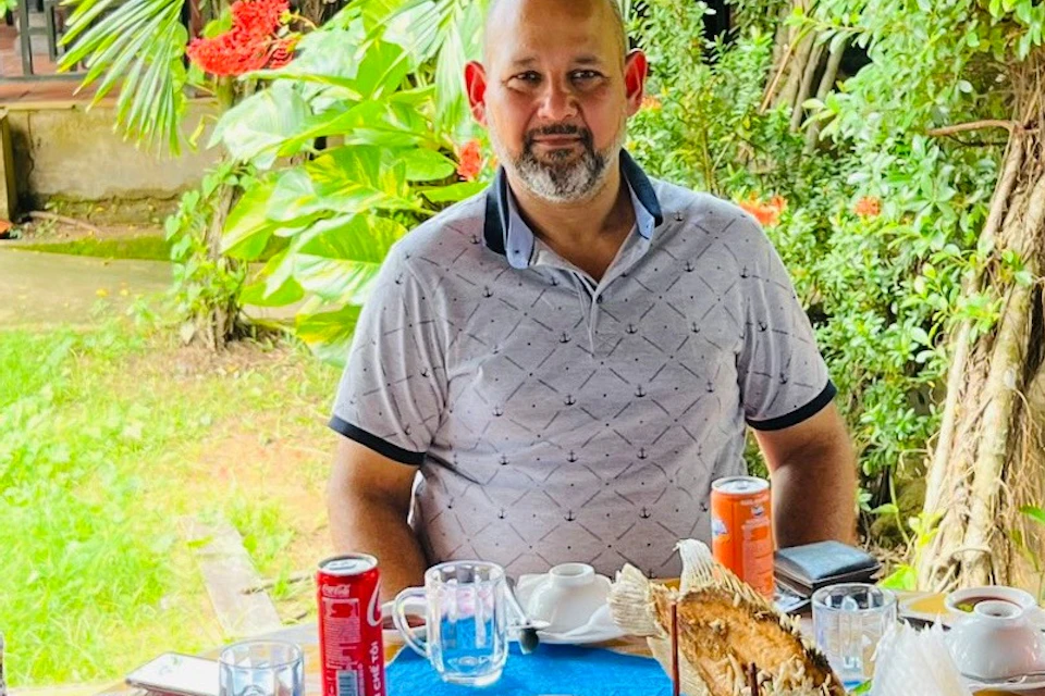 Three people seated at an outdoor table with food and drinks, surrounded by lush greenery. The tablecloth is yellow.