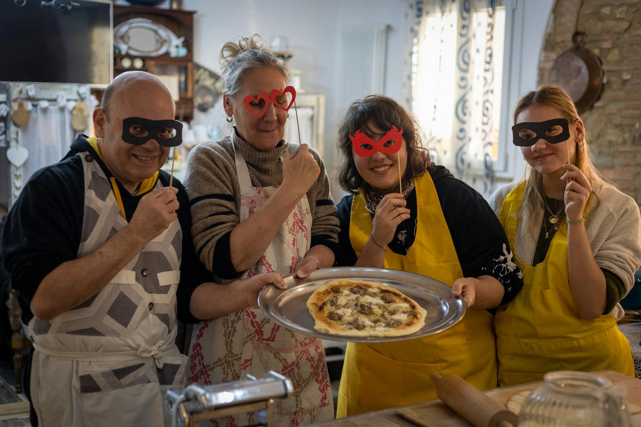 Tres persones amb davantal, sostenint màscares; una dona presenta una pizza en una safata. Entorn de cuina amb il·luminació lluminosa.