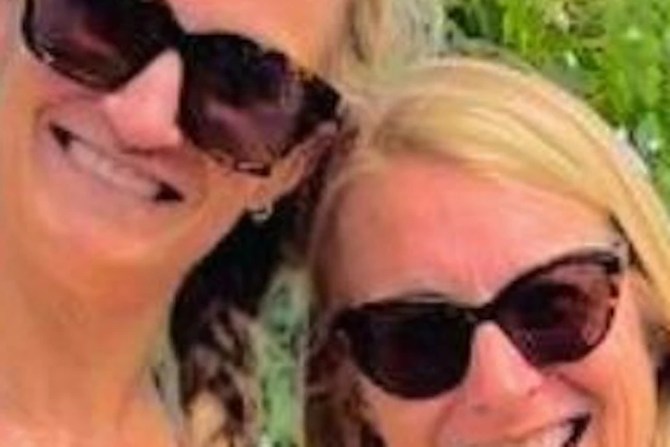 Two women smiling and posing in a vineyard, with grapevines in the foreground and a small red vehicle in the background.
