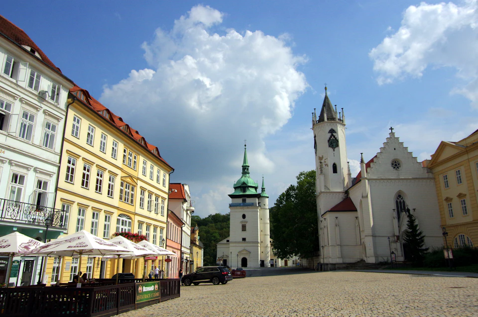 A cobblestone square with colorful buildings, two church towers, outdoor seating, and parked cars under a partly cloudy sky.