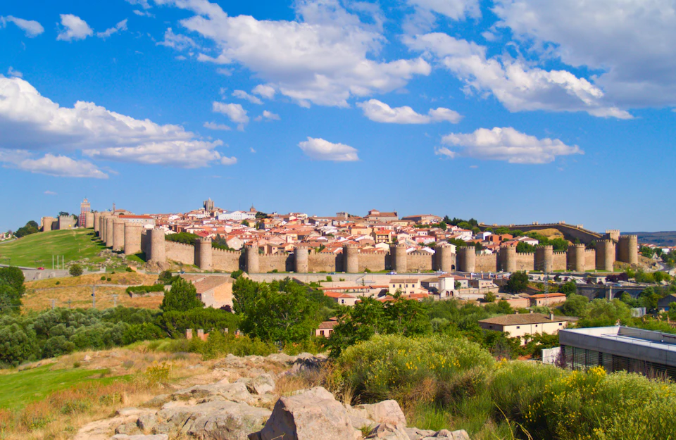 Scenic view of a historic walled city with multiple towers, surrounded by greenery, under a blue sky with scattered clouds.