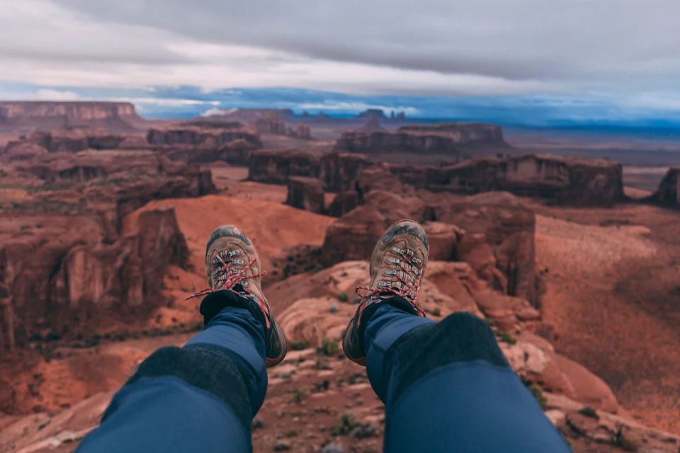 Hiking boots and legs in blue pants dangle over a vast desert canyon landscape with rock formations under a cloudy sky.