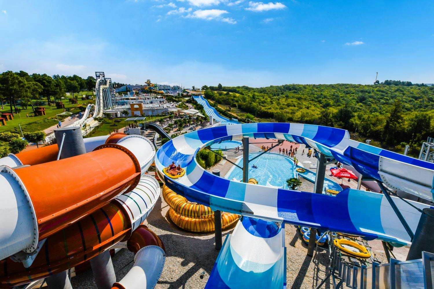 Farbenfrohe Wasserrutschen und Schwimmbecken in einem belebten Wasserpark vor der Kulisse grüner Hügel unter einem strahlend blauen Himmel.