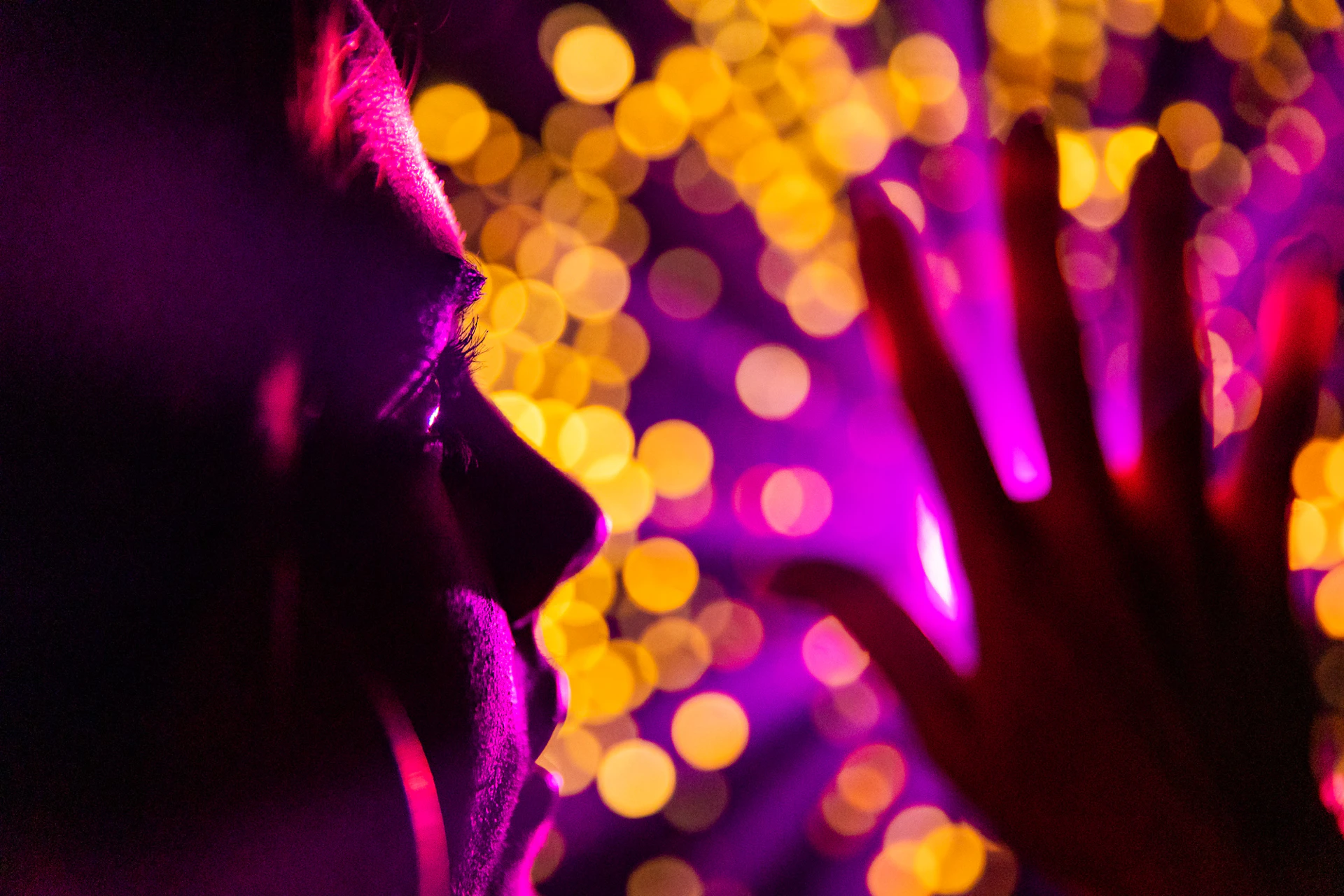 Silhouette of a person's face and hand against a background of vibrant yellow and purple bokeh lights.