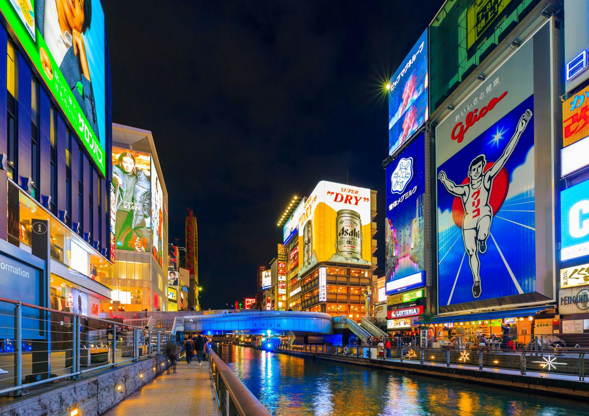 A vibrant night scene of a city canal with illuminated billboards and a pedestrian bridge in the background.