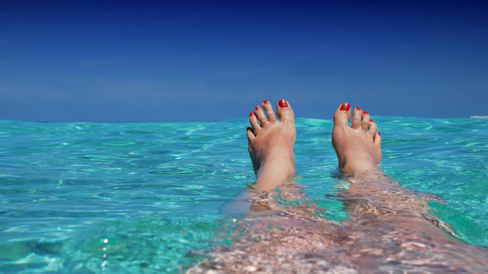 Feet with red toenails floating in clear turquoise water against a blue sky backdrop.