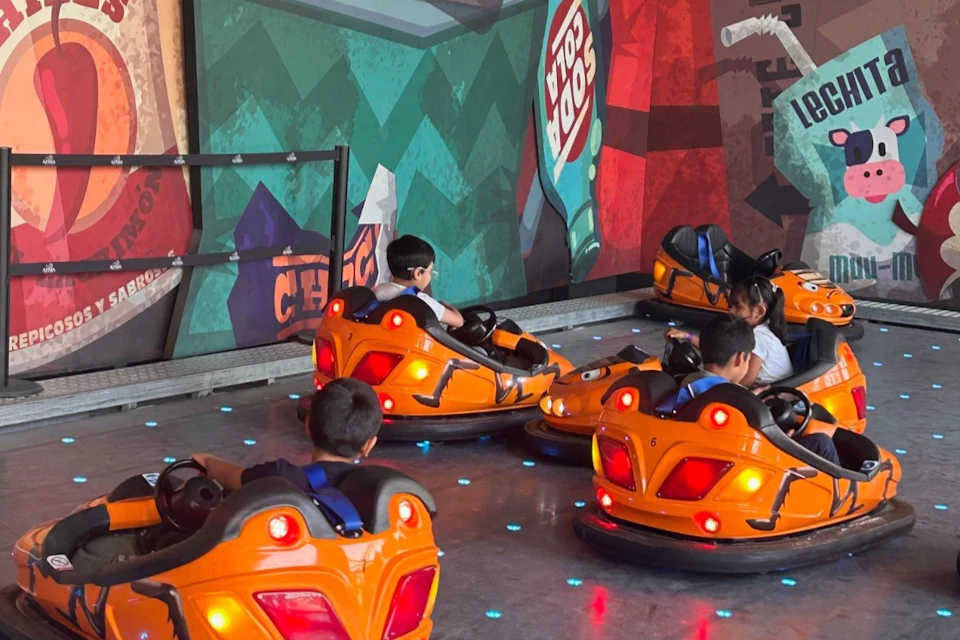 Children ride in orange bumper cars on a reflective floor with colorful mural backgrounds.