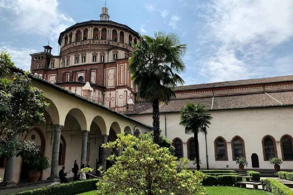 Courtyard with arched walkway, palm trees, and historic brick building with dome in the background under a cloudy sky.