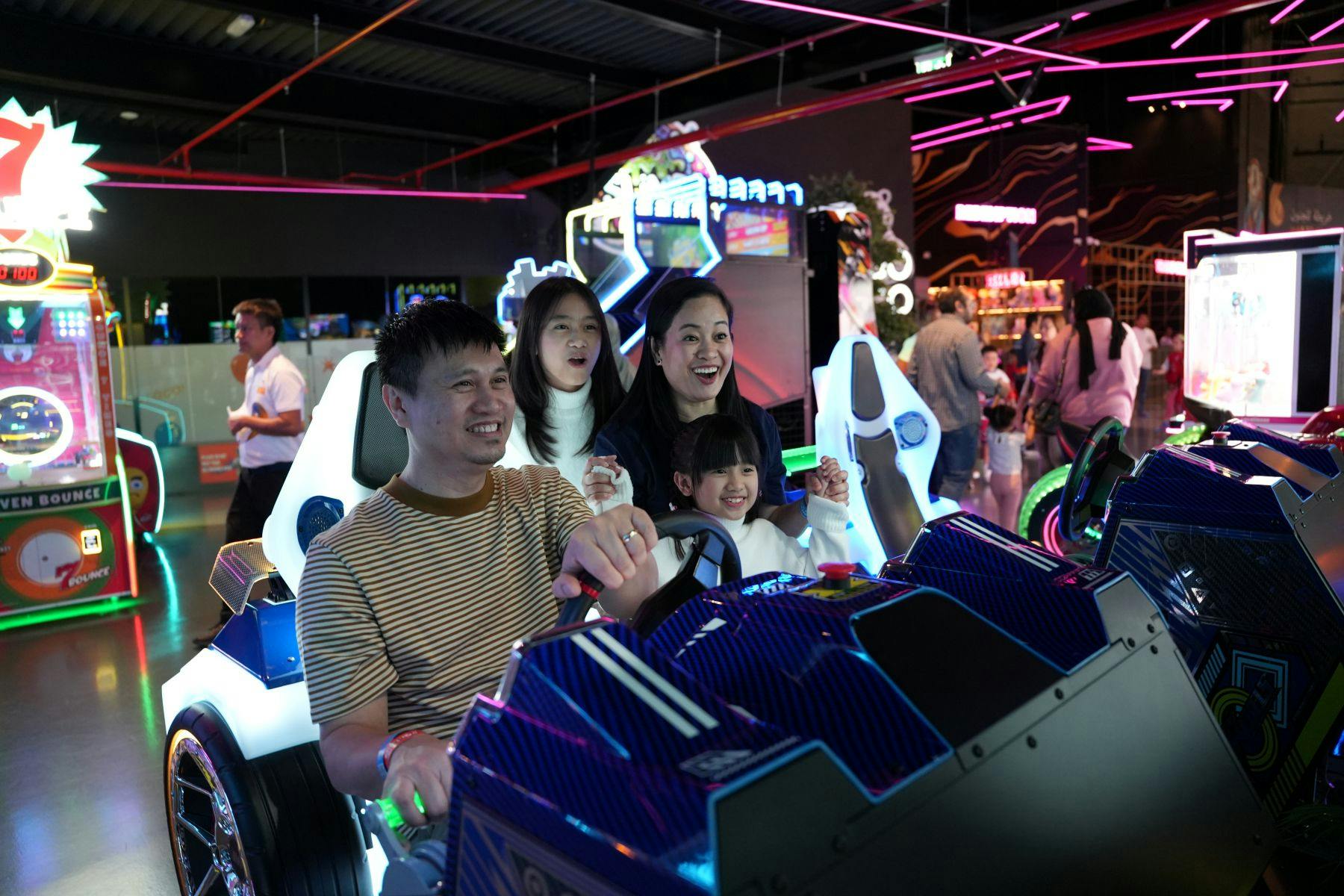Μια τετραμελής οικογένεια που απολαμβάνει ένα παιχνίδι οδήγησης arcade σε ένα έντονα φωτισμένο κέντρο τυχερών παιχνιδιών με πολύχρωμα φώτα νέον στο παρασκήνιο.