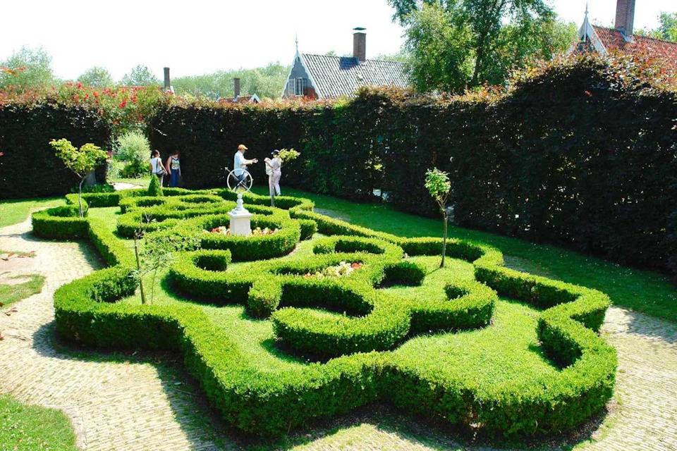 Garden with a well-manicured hedge maze, a white statue, several people exploring, and buildings in the background.