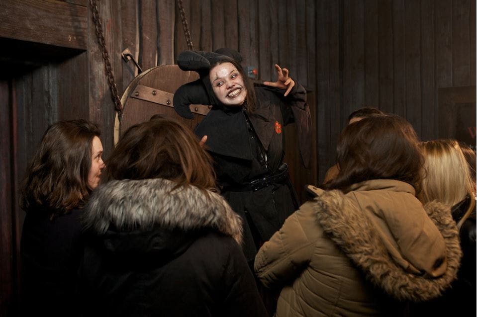 A person in Gothic makeup and attire poses dramatically in front of three people with winter coats inside a dimly lit room.