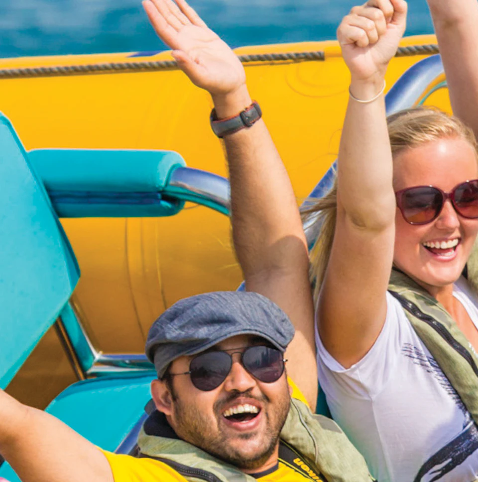 A group of people wearing life jackets, raising their hands, enjoying a ride on a yellow speedboat on the ocean under a clear blue sky.