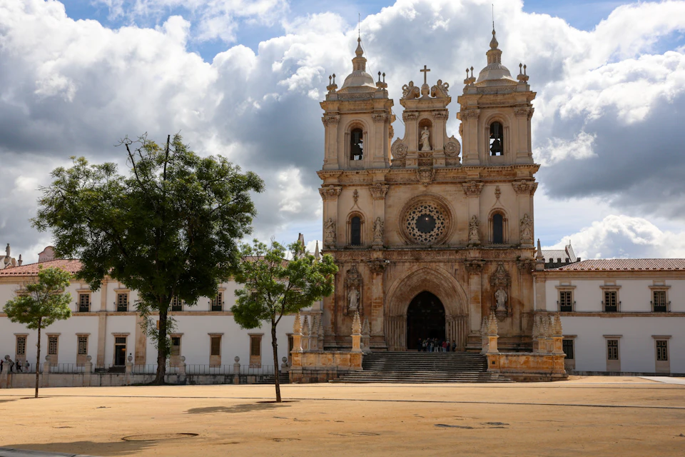A historic cathedral with twin bell towers and a large rose window, surrounded by a square and trees under a cloudy sky.