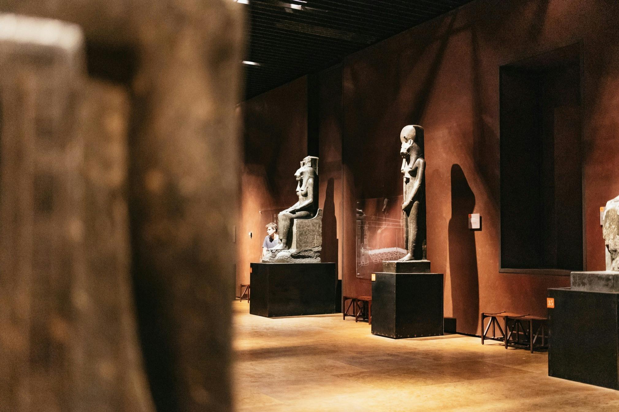 A museum gallery featuring ancient statues displayed on pedestals with soft lighting and warm-colored walls.