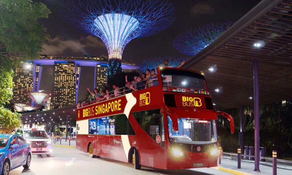 A red double-decker tour bus labeled 'Big Bus Singapore' with passengers on the top deck, set against a lit nighttime cityscape.