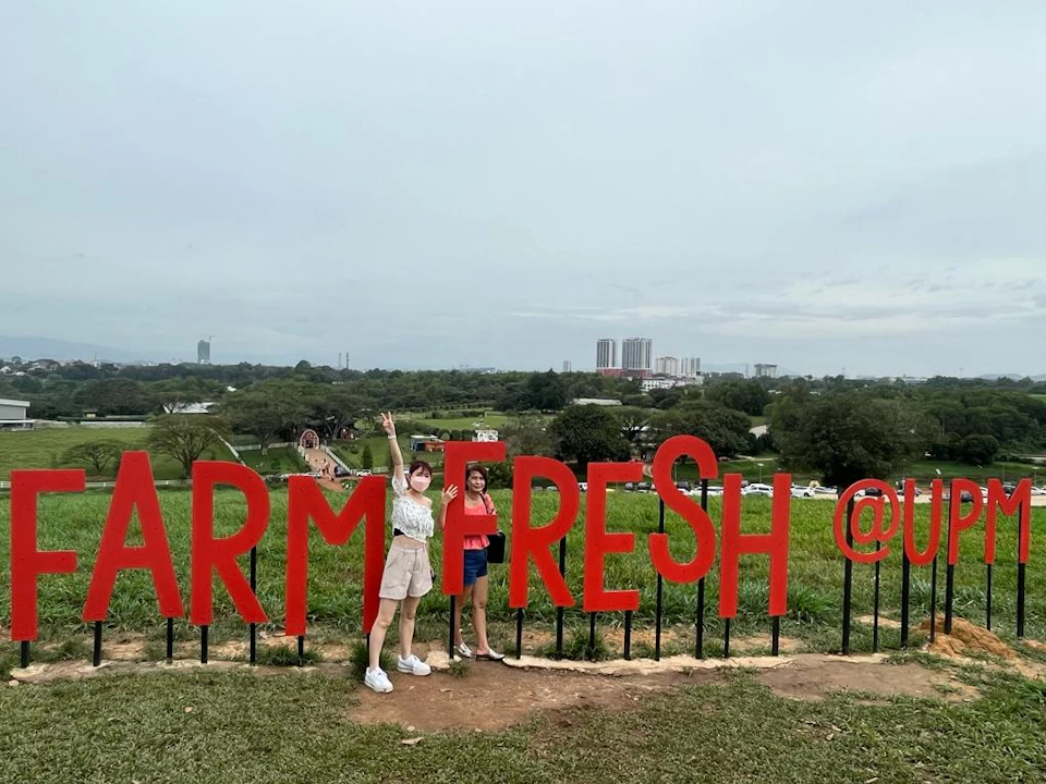 Two people stand next to large red letters spelling "FARM FRESH" in a grassy area with a city skyline in the background.
