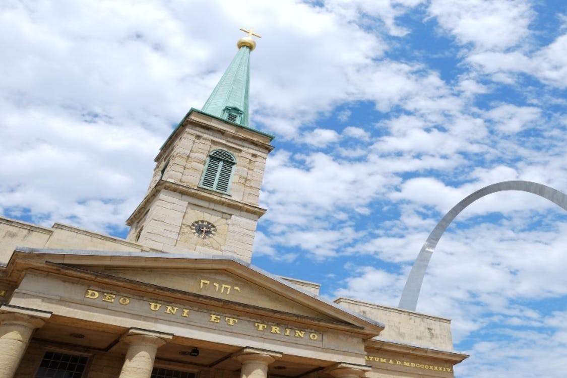 Μια ιστορική εκκλησία με πράσινο καμπαναριό, ρολόι και σταυρό κάτω από έναν εν μέρει συννεφιασμένο ουρανό, με ορατό μέρος μιας σύγχρονης δομής αψίδας.