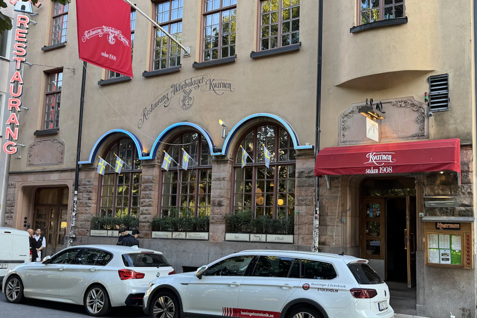 A historic restaurant with arched windows, Swedish flags, and parked cars in front. Red awning displaying the name.