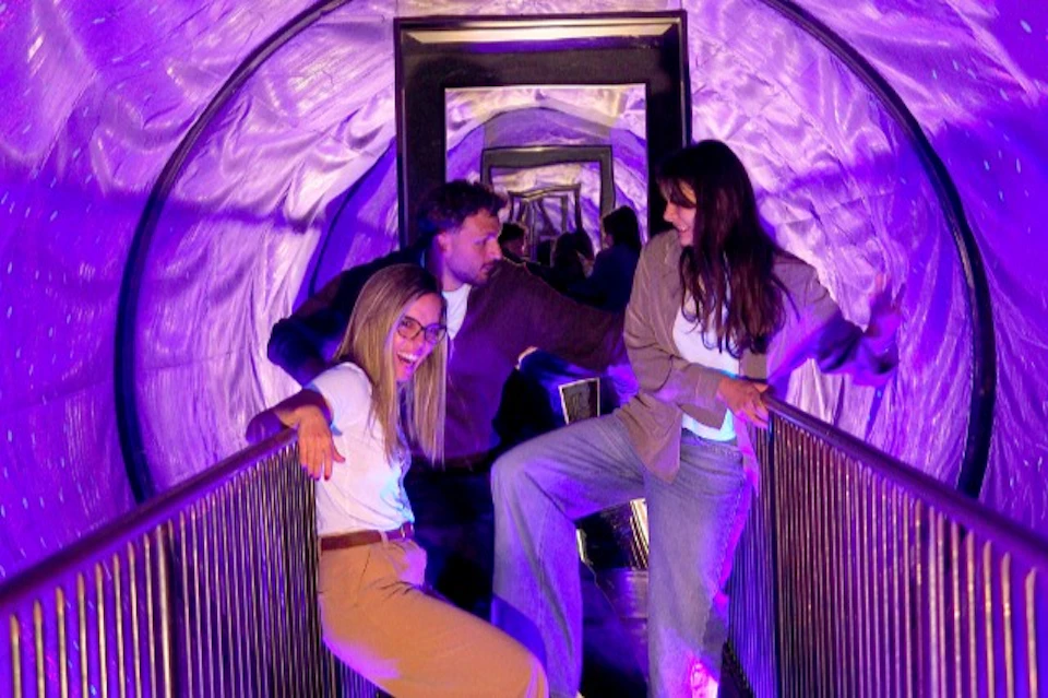 Three people stand on a bridge in a tunnel with purple lighting and mirrored walls, posing and interacting playfully.