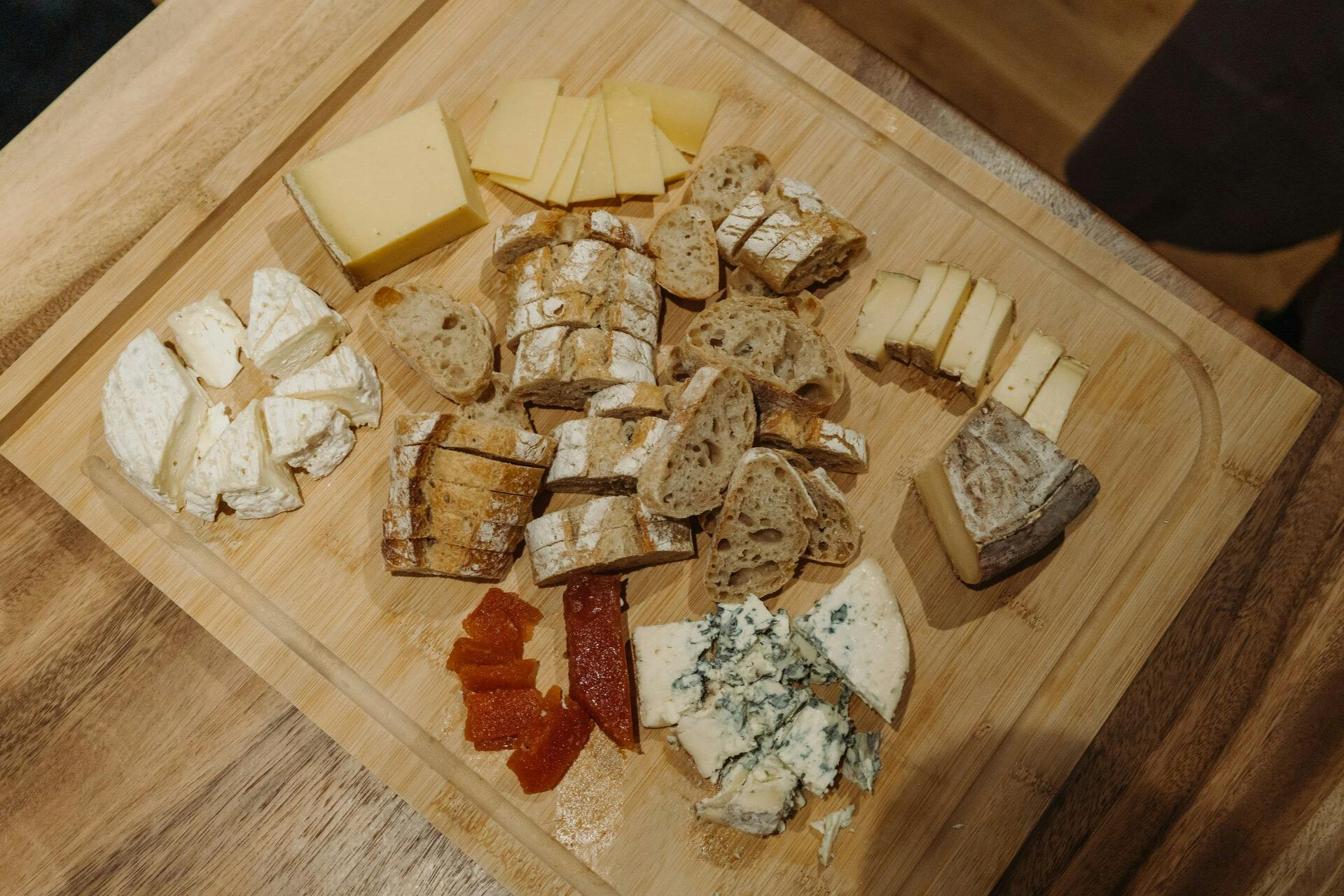 A wooden board with assorted cheeses, sliced bread, and small portions of fruit preserves arranged for serving.