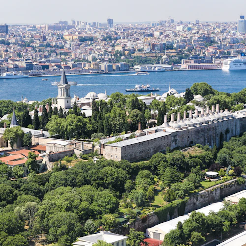 Historiskt palatskomplex med torn och kupoler omgivna av frodig grönska, nära en flod med en stadsbild i bakgrunden.
