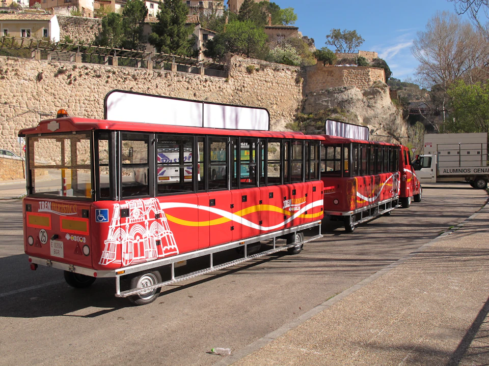 A red tourist train with an accessible entrance symbol parked beside a stone wall and trees on a sunny day.