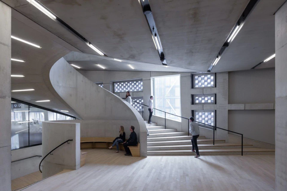 A modern interior space with concrete walls, a staircase, and people walking or sitting on benches under bright lights.