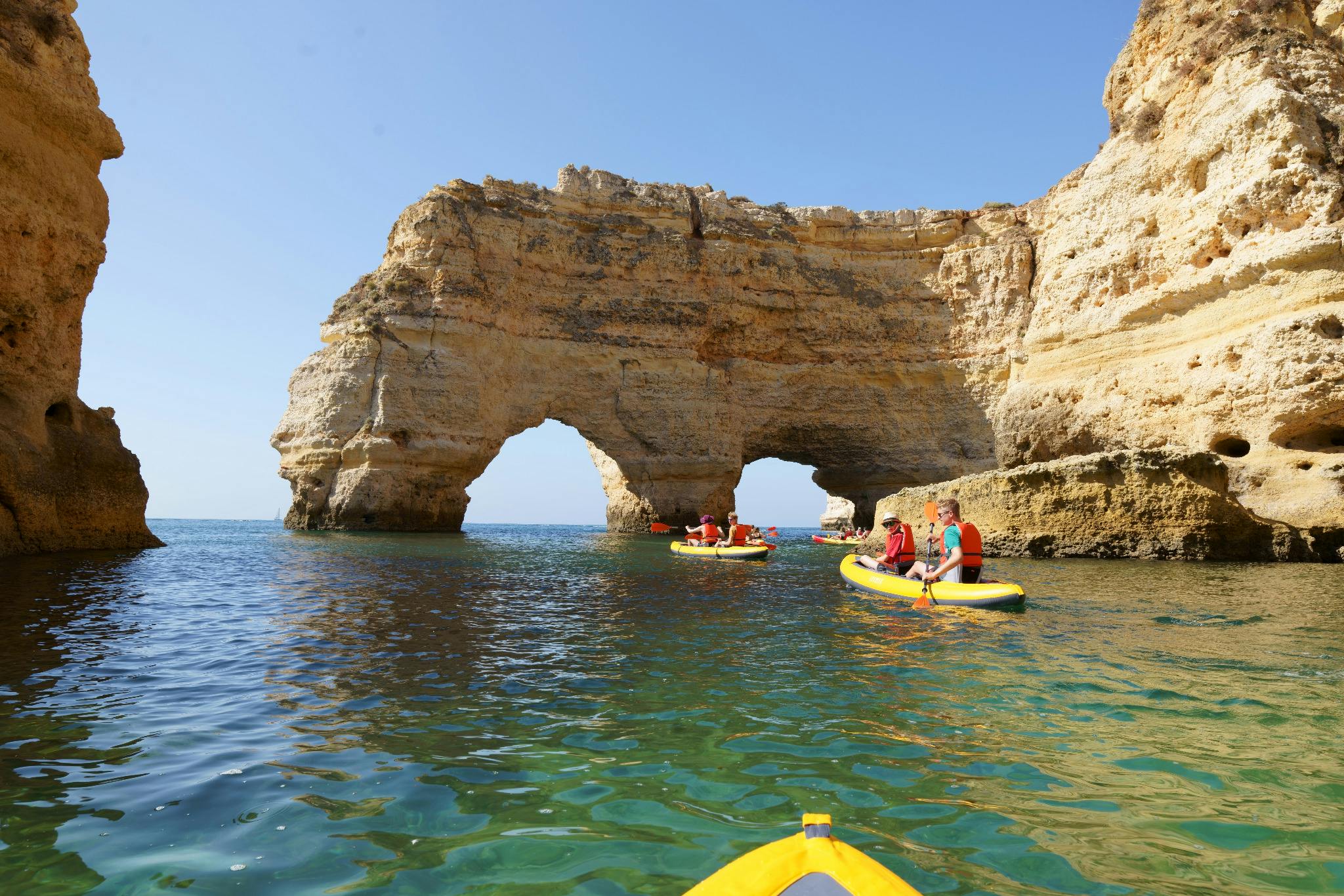 Kajakáři proplouvají průzračnou vodou kolem velkých skalních oblouků pod jasně modrou oblohou.
