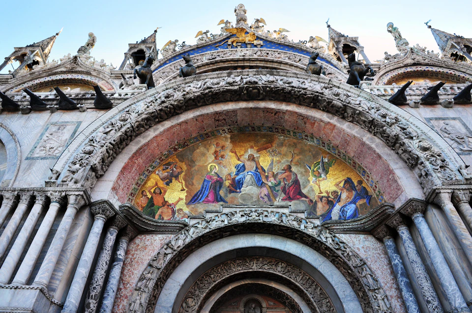 Intricate arched facade of a historical building with detailed carvings and a colorful mosaic depicting religious figures.