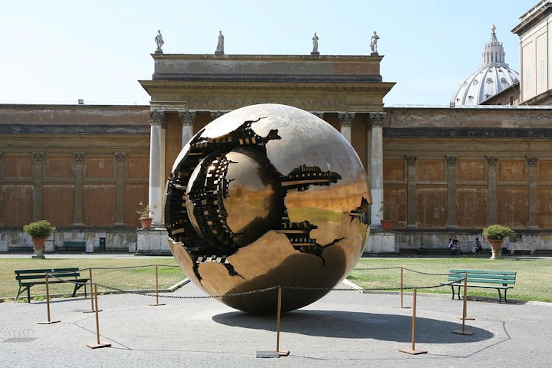 A large bronze spherical sculpture with intricate inner designs stands in front of a classical building with columns and statues.