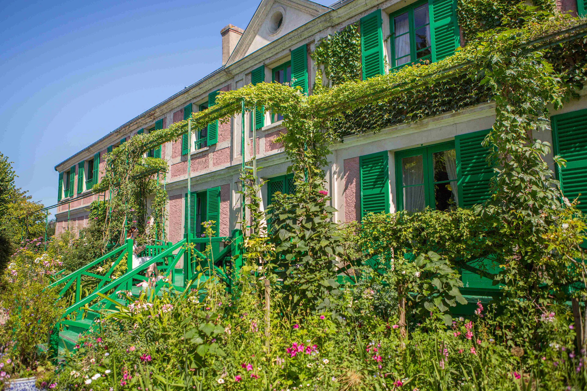 A building with green shutters and ivy-covered walls, surrounded by lush greenery and colorful flowers under a blue sky.