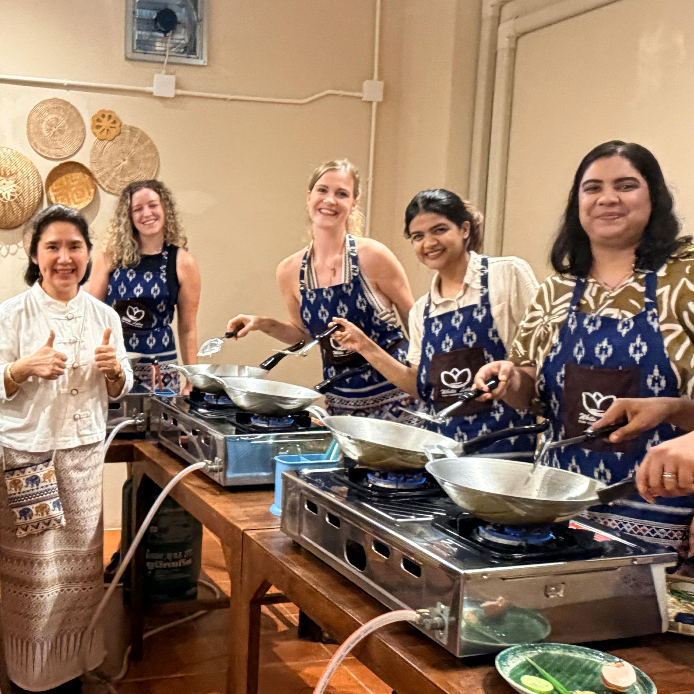 A group of women wearing aprons stand at a cooking station with woks on portable stoves, smiling at the camera.