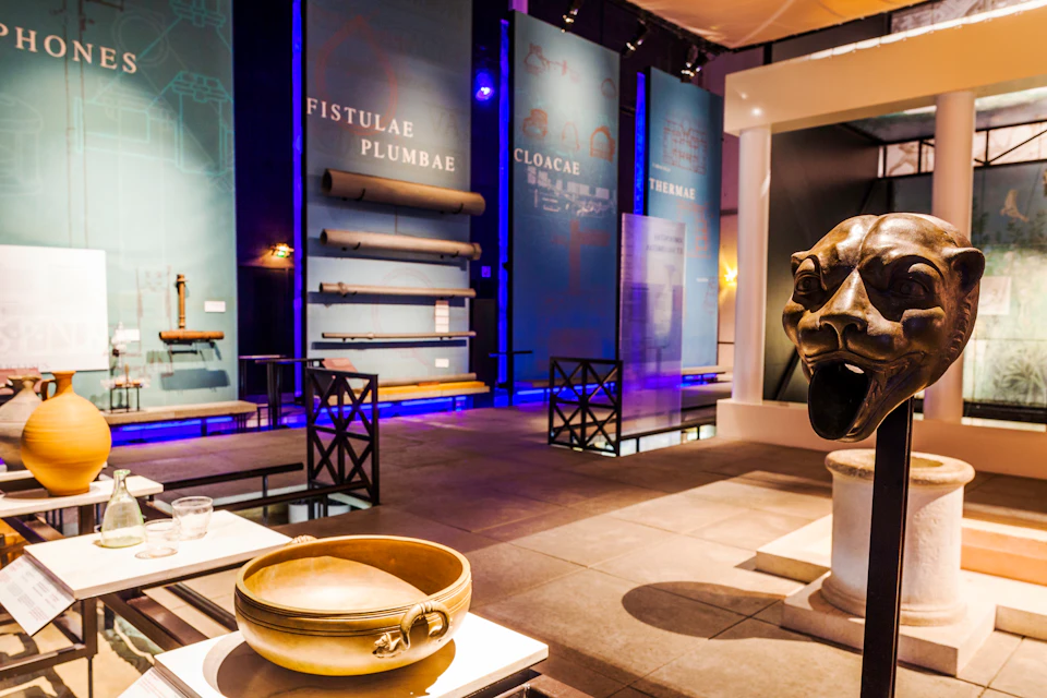 Museum exhibit featuring ancient Roman plumbing artifacts, including pipes and basins. Large lion head sculpture in the foreground.