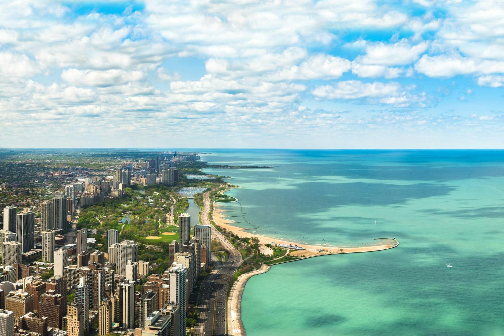 Aerial view of a city with tall buildings along a coastline, featuring beaches, green parks, and a vast blue lake beneath a partly cloudy sky.