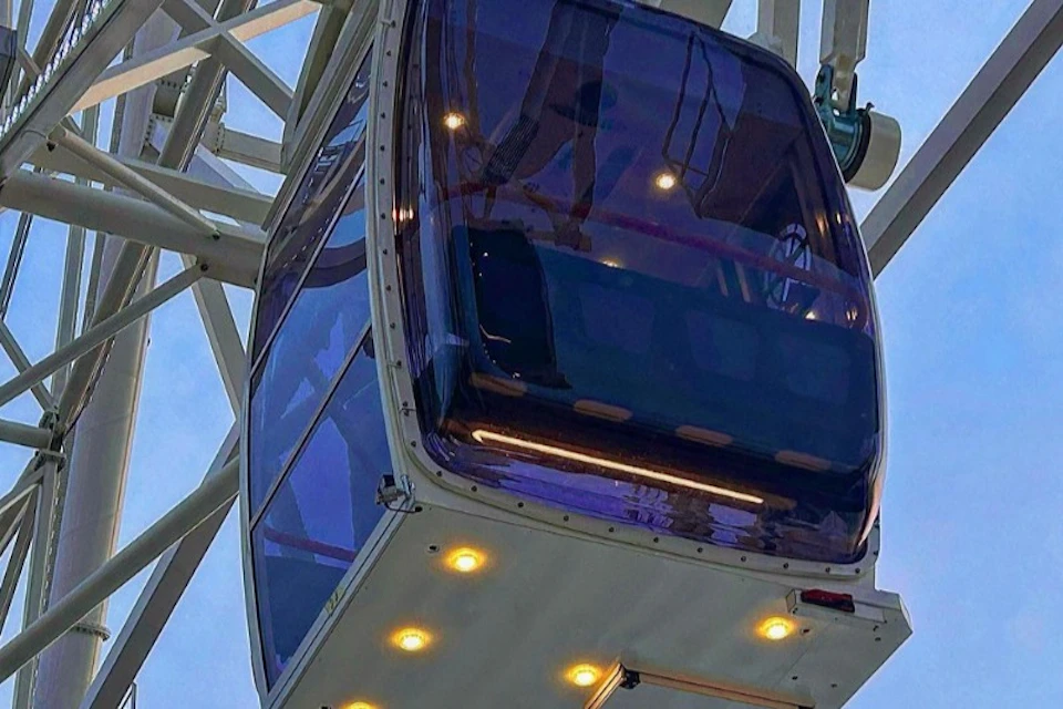 A gondola on a Ferris wheel with reflective tinted windows and illuminated lights underneath, against a clear sky.