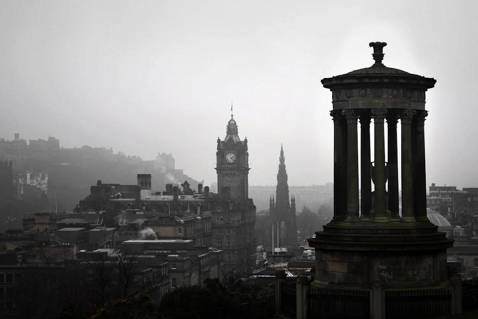 Foggy cityscape with historic buildings, including a clock tower and a monument with columns in the foreground.
