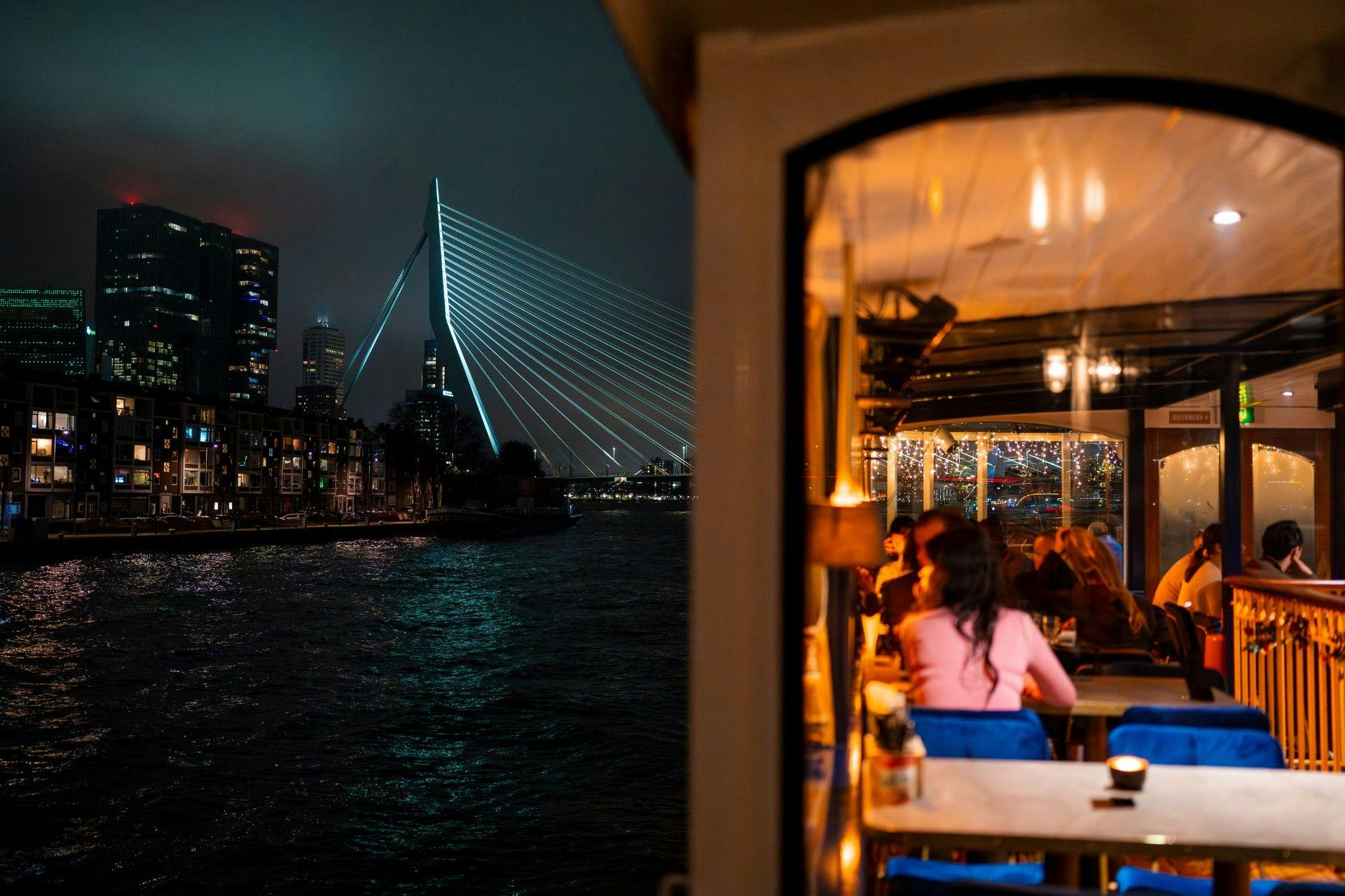 Nattvy av en upplyst bro över vatten, med en starkt upplyst restaurangterrass i förgrunden.