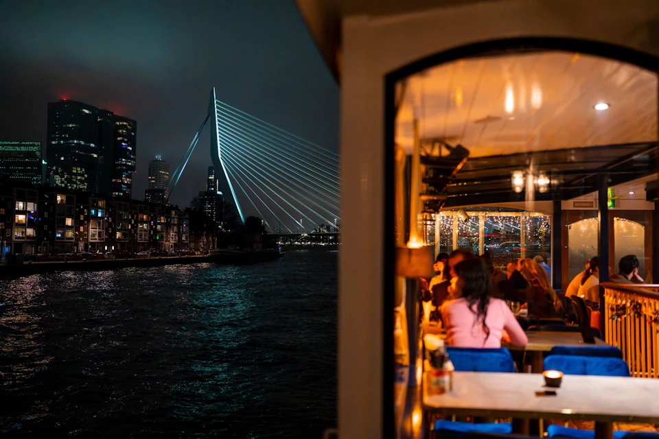 Nighttime view of a lit bridge over water, with a brightly illuminated restaurant terrace in the foreground.