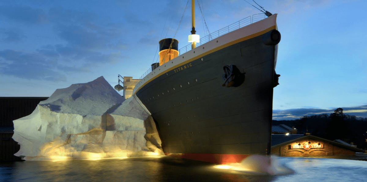 A large Titanic replica with an iceberg structure beside it, against a twilight sky. &#34;White Star&#34; is visible on a nearby building.